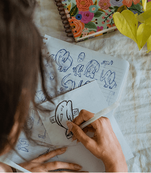Person drawing on a piece of paper with a colorful notebook and plant in the background