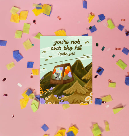 Greeting card with a mountain scene and a little dog riding a gondola. At the top the text says 'you're not over the hill (quite yet)'.