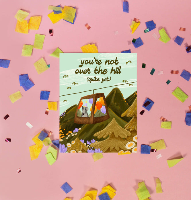 Greeting card with a mountain scene and a little dog riding a gondola. At the top the text says 'you're not over the hill (quite yet)'.