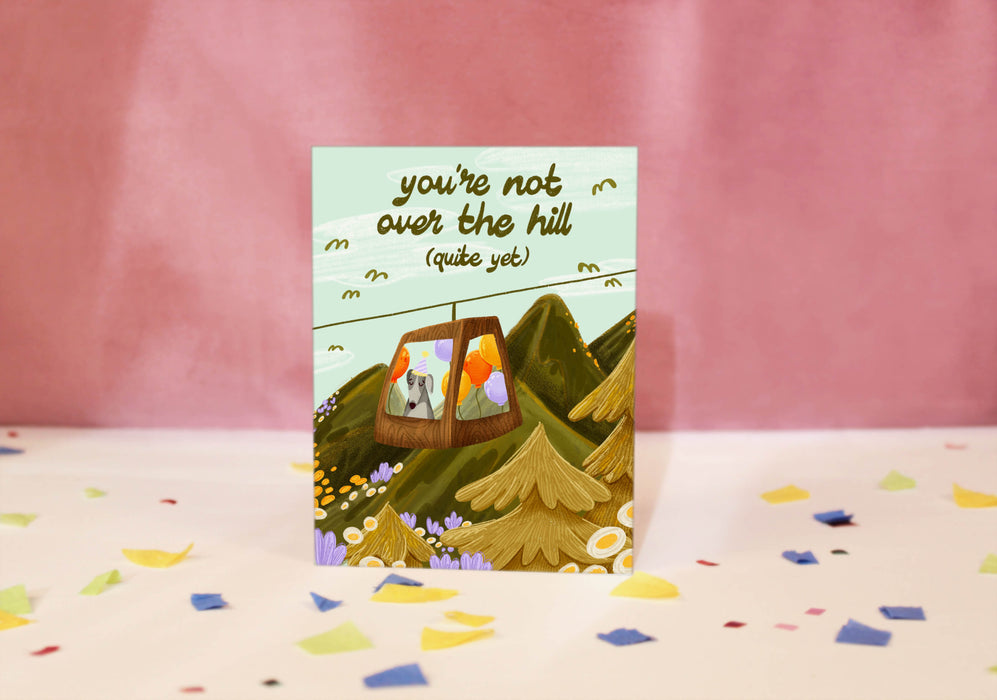Greeting card with a mountain scene and a little dog riding a gondola. At the top the text says 'you're not over the hill (quite yet)'.