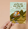 Greeting card with a mountain scene and a little dog riding a gondola. At the top the text says 'you're not over the hill (quite yet)'.