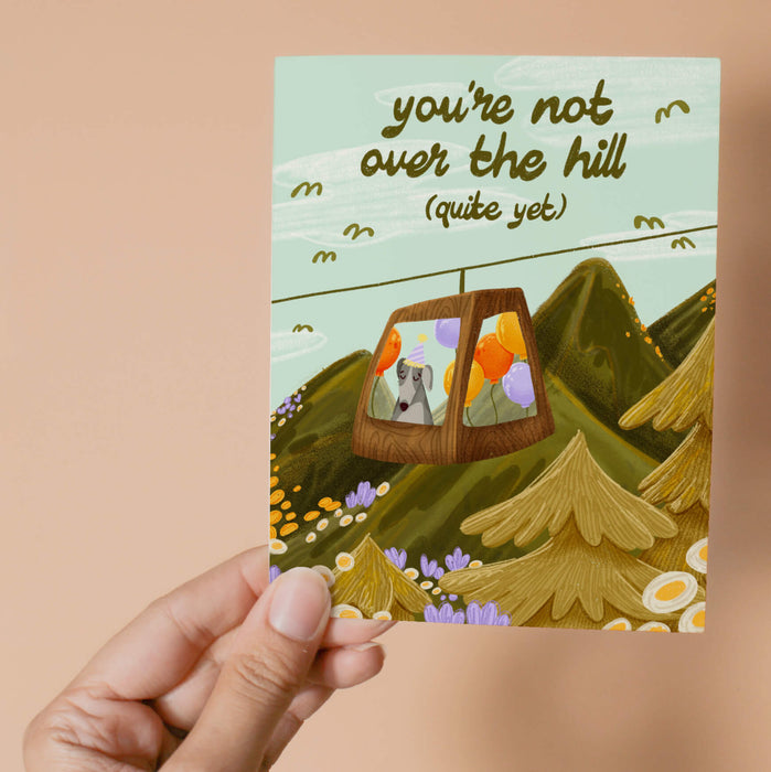 Greeting card with a mountain scene and a little dog riding a gondola. At the top the text says 'you're not over the hill (quite yet)'.