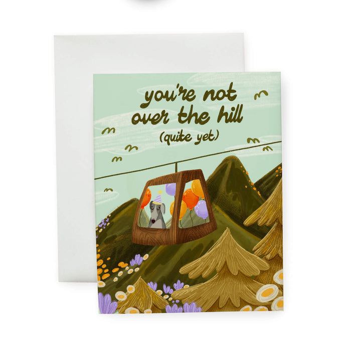 Greeting card with a mountain scene and a little dog riding a gondola. At the top the text says 'you're not over the hill (quite yet)'.