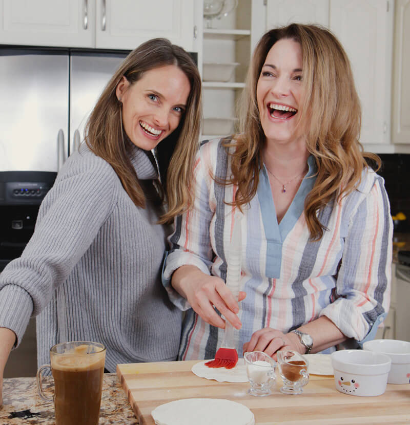 Two women in a kitchen preparing food, with a coffee cup and cutting board in the foreground.