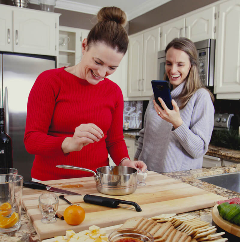 Two women in a kitchen, one cooking and the other using a phone.