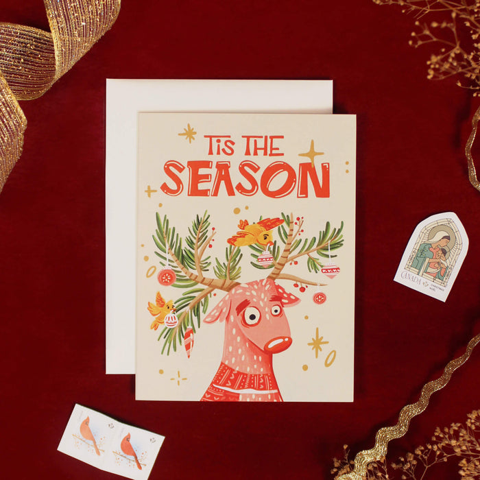 Christmas card with a reindeer illustration and 'Tis the Season' text on a red background. The antlers are tree branches are birds are decorating them with ornaments,
