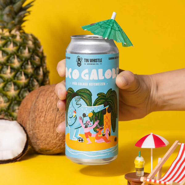 Hand holding a can of 'Coco Galore' beer with a pineapple, coconut, and beach-themed decorations on a yellow background.