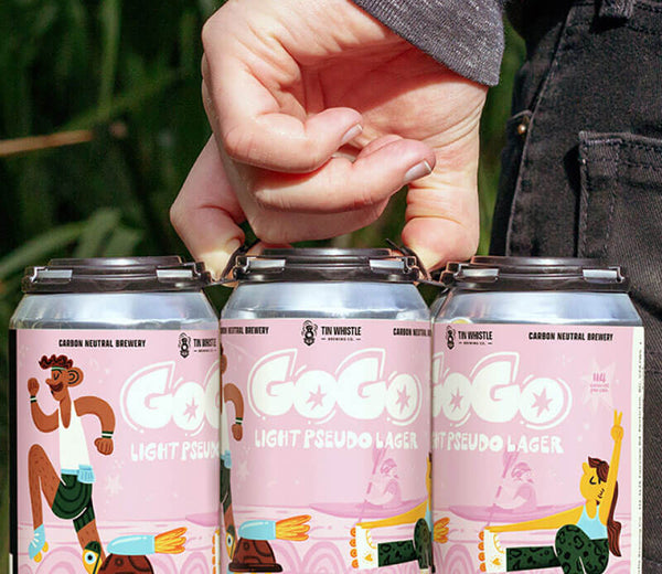 Three cans of GoGo Light Pseudo Lager with a person's hand holding them, set against a natural background.
