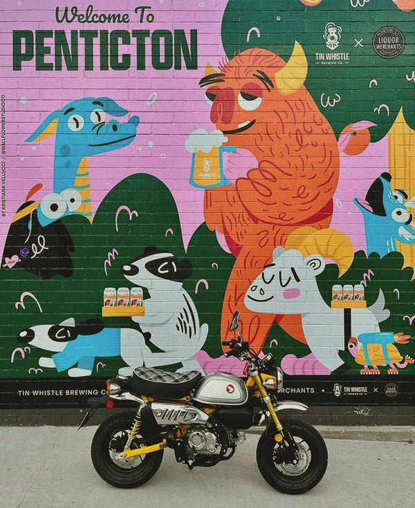 Motorcycle parked in front of a colorful mural with cartoon animals and 'Welcome to Penticton' text.
