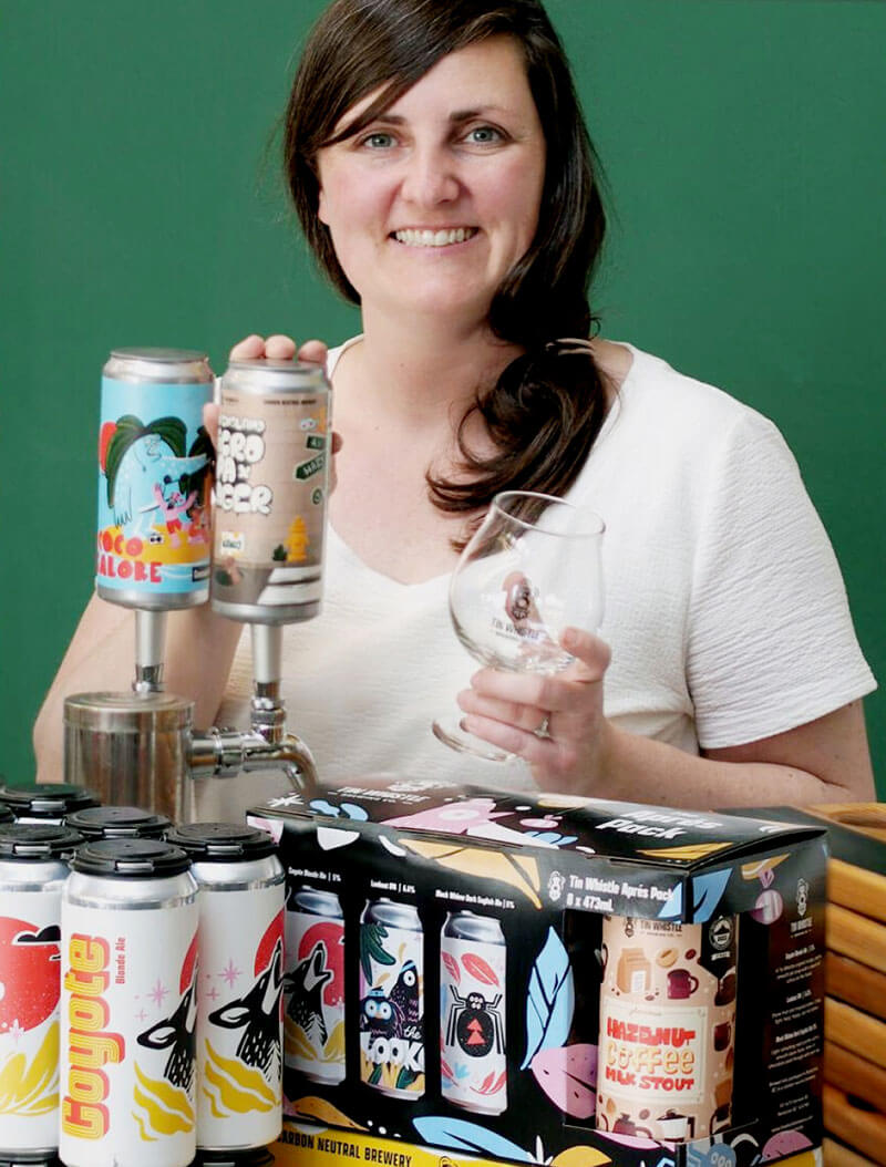 Woman holding beer cans and a glass with a package of beer on the table in front of her.