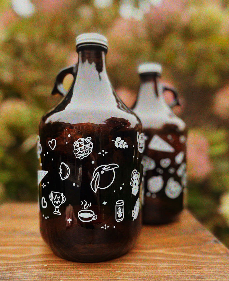 Two brown glass growlers with white illustrated icons on a wooden surface with a blurred natural background